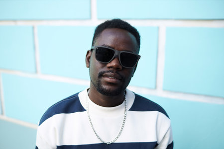 Colorful front view portrait of funky African American man wearing sunglasses and looking at camera against blue brick wall, copy spaceの写真素材