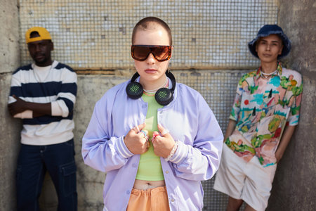 Portrait of three young people wearing trendy outfits standing in urban setting with bald young woman in foreground.の写真素材