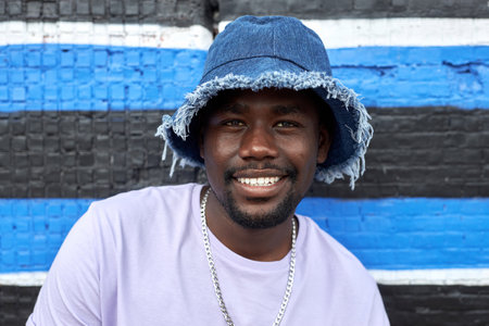 Front view portrait of smiling young black man wearing jeans hat and looking at camera while posing in graphic urban setting against brick wallの写真素材