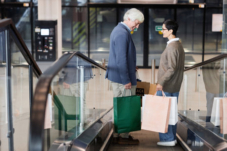 Side view of senior couple holding shopping bags standing on escalator in shopping mall, copy spaceの写真素材