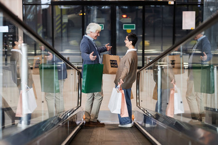 Side view of happy mature couple holding shopping bags standing on escalator in shopping mall, copy spaceの写真素材