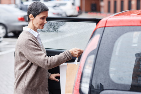 Side view portrait of mature woman putting shopping bags in car outdoors in parking lot, copy spaceの写真素材