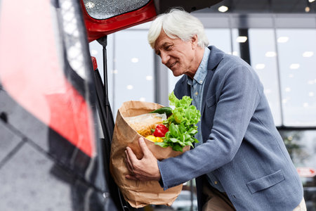 Side view portrait of smiling senior man putting grocery bags in car trunk outdoors, copy spaceの写真素材