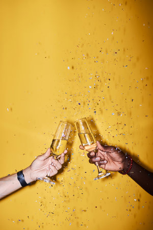 Close up of two people clinking champagne glasses against vibrant yellow background with confetti popの写真素材