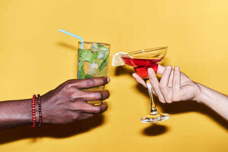 Close up of multiethnic young couple holding two cocktails against vibrant yellow background