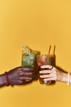 Close up of couple hands clinking cocktails against vibrant yellow background in celebration, copy spaceの写真素材