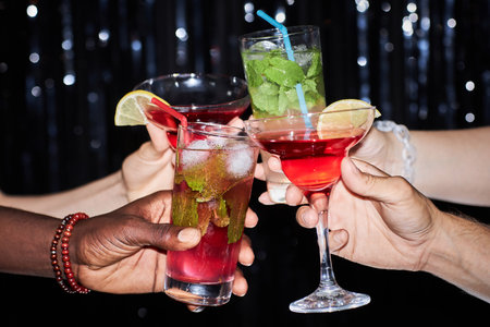 Close up of group of friends holding cocktails and toasting against glittering party background,...