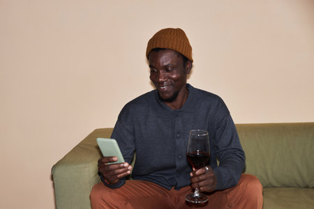 Minimal portrait of young Black man using smartphone and drinking red wine, shot with flash, copy spaceの写真素材