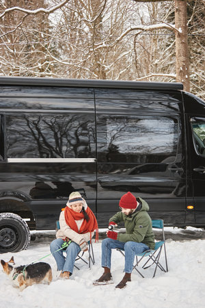 Vertical portrait of young couple enjoying hot coffee outdoors while travelling with trailer van in winterの写真素材