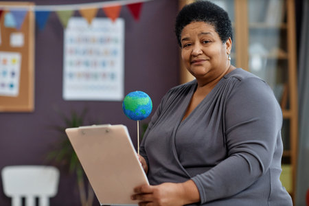 Portrait of kind black woman as female teacher holding clipboard in preschool and looking at camera, copy spaceの写真素材