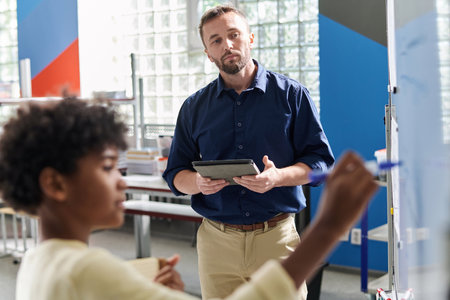 Serious teacher looking at kid writing formulas on whiteboardの写真素材