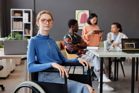 Portrait of young woman using wheelchair looking at camera in modern office with team in background, copy spaceの写真素材