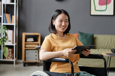 Portrait of Asian young woman using wheelchair smiling at camera and holding tablet in office, copy spaceの写真素材