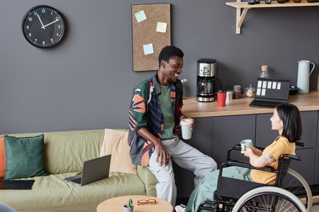 Side view portrait of young woman using wheelchair in office and chatting with smiling colleague during coffee break, copy spaceの写真素材
