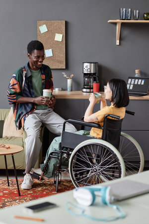Vertical full length portrait of young woman in wheelchair enjoying chat with colleague during coffee breakの写真素材