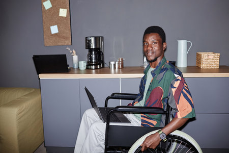 Portrait of one young Black man using wheelchair and smiling at camera in modern office lounge, shot with flash, copy spaceの写真素材