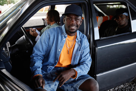 Young smiling male hip hop dancer in black cap, denim jacket and shorts sitting on front seat of car...
