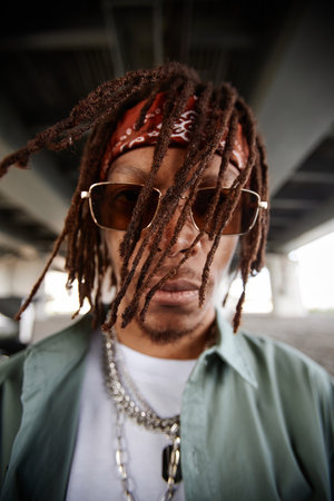 African American guy with dreadlocks looking at camera through sunglasses while standing in urban...