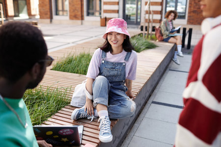 Portrait of carefree young Asian girl talking to group of friends on campus outdoors and wearing fun...