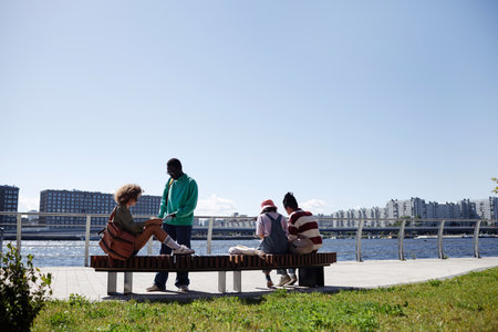 Back view at diverse group of young people relaxing outdoors by river against city skyline, copy spaceの写真素材