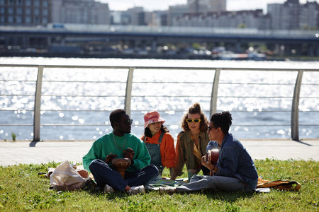 Wide angle view of young people enjoying picnic on grass outdoors with city skyline in background, copy spaceの写真素材