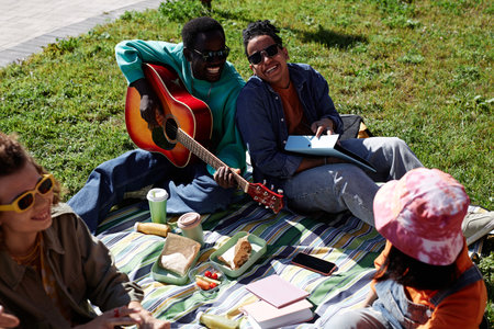 High angle view at friends enjoying picnic on grass in sunlight and playing guitar, copy spaceの写真素材