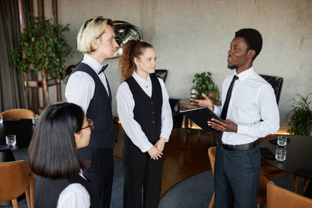 Side view portrait of young Black man as restaurant manager talking to servers wearing classic...