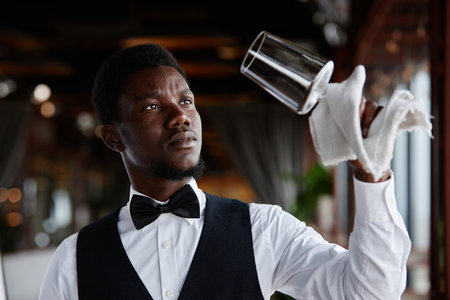 Portrait of Black young man as elegant waiter polishing glasses in luxury restaurant, copy spaceの写真素材