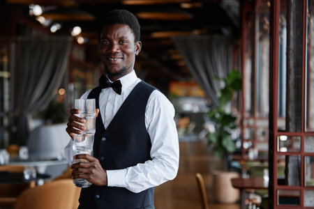 Waist up portrait of young Black man as elegant server holding glasses and smiling at camera in...