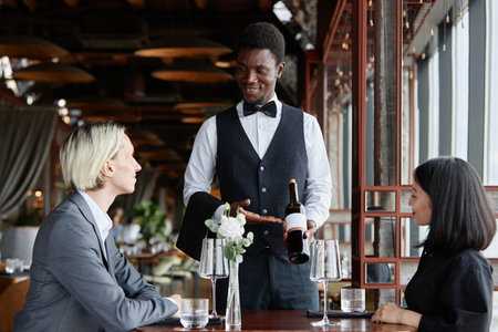 Portrait of Black young man as server taking orders from couple in luxury restaurant and presenting bottle of red wine, copy spaceの写真素材