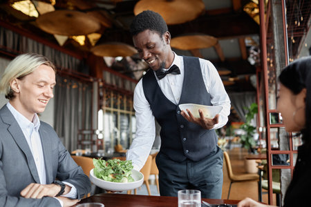 Portrait of young Black man as server bringing food on tray to couple enjoying dinner in luxury restaurantの写真素材