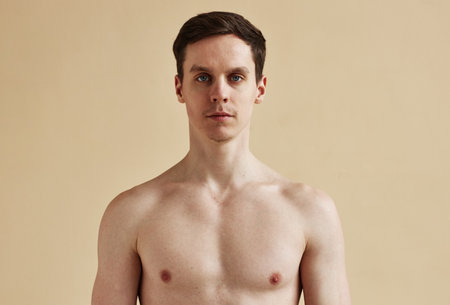 Minimal portrait of shirtless young man looking at camera with blue eyes against pale background,...