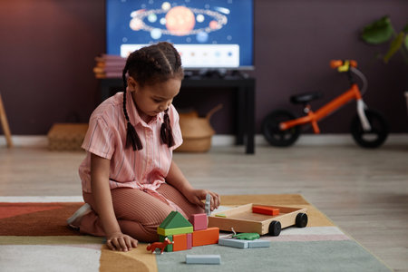 Full length portrait of little Black girl playing with wooden toys while sitting on soft carpet in...