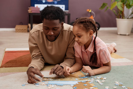 Portrait of Black little girl solving jigsaw puzzle while laying on floor with father and playing together at homeの写真素材