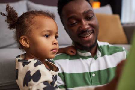 Side view portrait of cute Black girl looking at pictures while reading book with father at home and enjoying family timeの写真素材