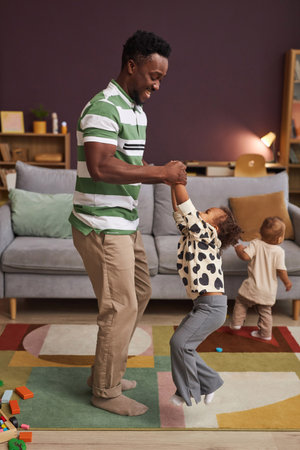Vertical full length portrait of happy Black father dancing with cute little girl at home in living roomの写真素材