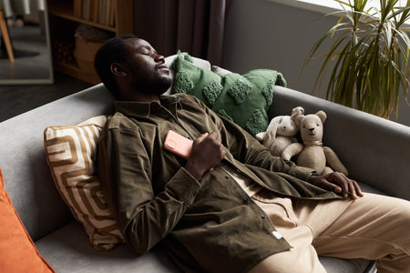 High angle portrait of Black adult man falling asleep on couch at home with smartphone on chest, copy spaceの写真素材