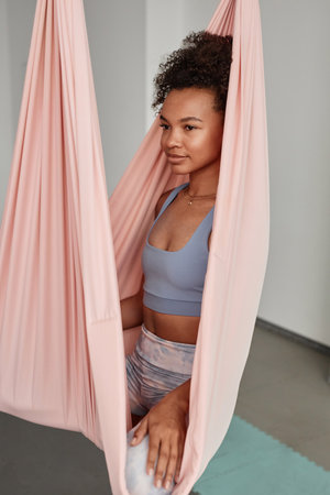Vertical portrait of young African American woman sitting in pink hammock enjoying aerial yoga in studioの写真素材