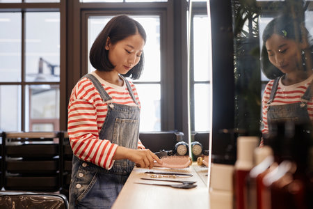 Side view portrait of young Asian hairstylist setting up tools and smiling while enjoying work in beauty salon, copy spaceの写真素材