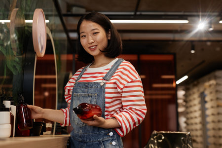 Waist up portrait of smiling Asian hairstylist looking at camera in beauty salon and holding shampoo bottles, copy spaceの写真素材