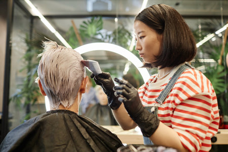 Side view portrait of young Asian hairstylist applying hair dye or bleach to male client in beauty salonの写真素材