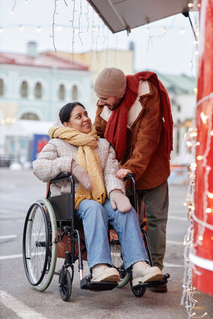 Full length portrait of young woman using wheelchair enjoying date with boyfriend outdoors in Christmas marketの写真素材