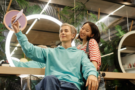 Portrait of smiling Asian man taking selfie in beauty salon with hairstylist, copy spaceの写真素材