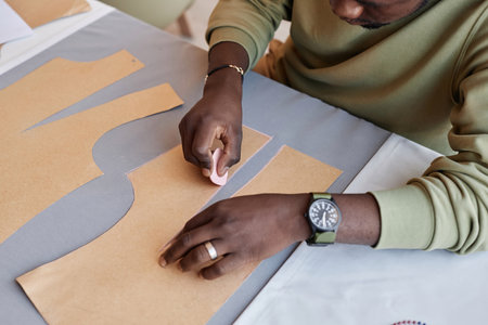 Young male tailor with piece of chalk in hand contouring paper patterns on grey textile while...