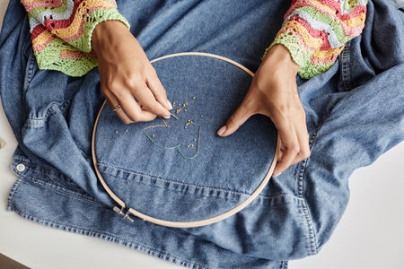 Above angle of hands of young creative woman making embroidery with beads on back of denim jacket...