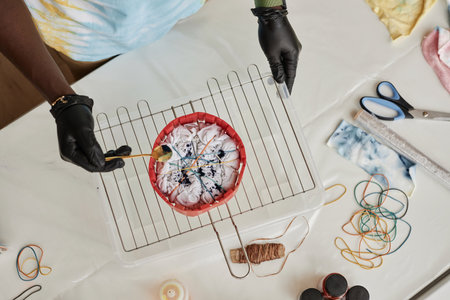 Above view of gloved hands of young man sprinkling powder paint on crumpled t-shirt bound with...