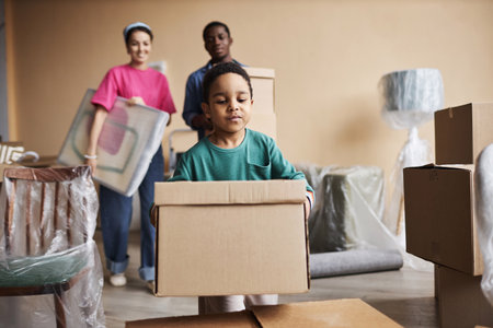 Cute little boy carrying packed cardboard box and putting it on top of another one while helping his...