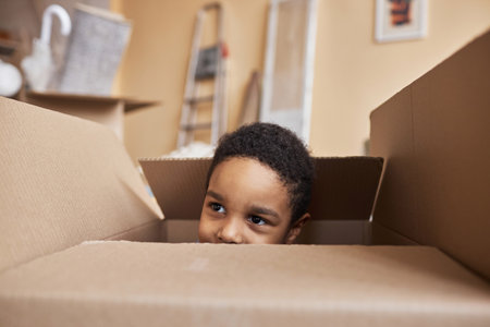 Face of cute little boy peeking out of big cardboard box and looking aside while playing and hiding from his parents during relocationの写真素材