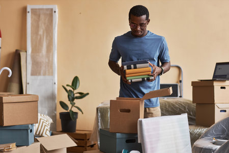 Young African American man with stack of books packing or unpacking boxes while standing in living room of new apartment or houseの写真素材