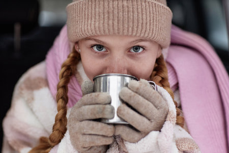 Focus on face of pretty pre-teenage girl in winter drinking hot tea and looking at camera while holding metallic mug in gloved handsの写真素材